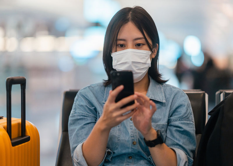 Woman seated in airport gate lounge checks her phone. She is wearing a mask to protect against COVID-19 transmission