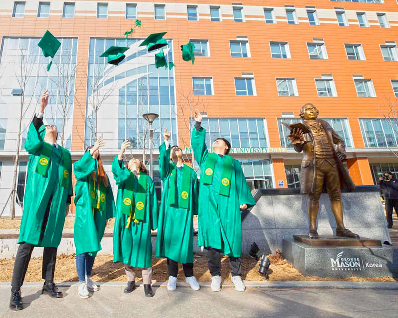 Students in graduation garb toss hats in the air in celebration of graduation at Mason Korea. They stand next to the Mason Korea statue of George Mason. 