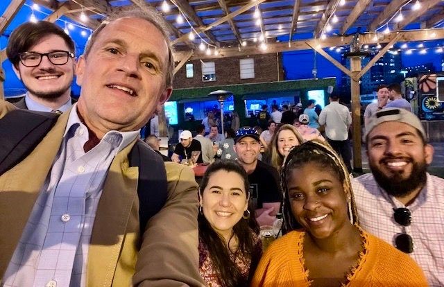 A man in a beige jacket takes a selfie surrounded by grad students.