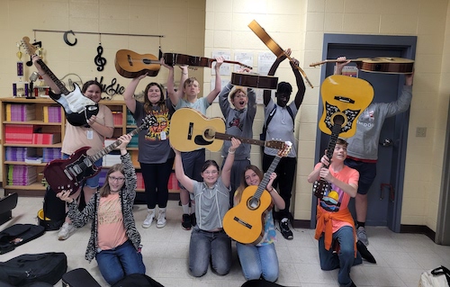 A group of grinning students hold up guitars for the camera, grinning. Many of the kids hold their guitars above their heads in exuberance.