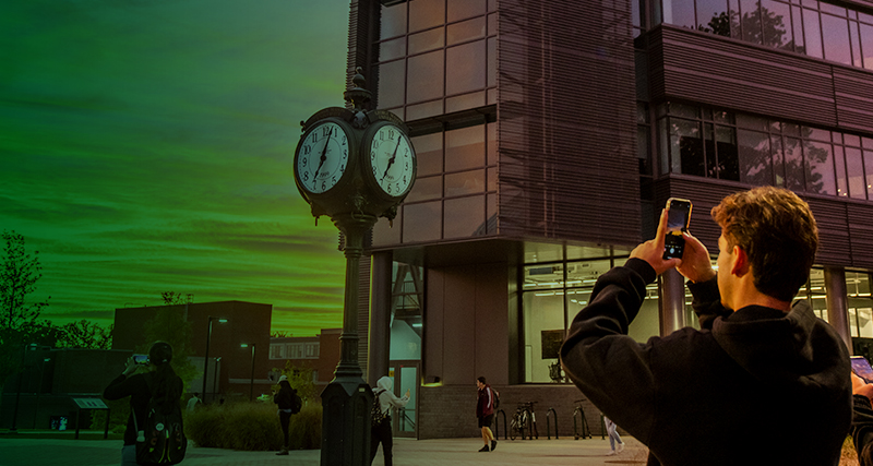 A student pauses in Wilkins Plaza to take a photo of the Mason clock during a beautiful sunset.