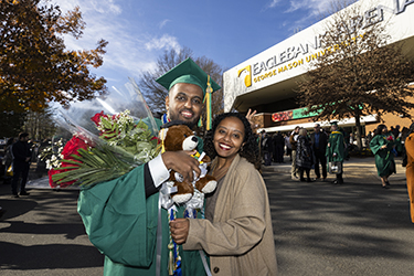 A graduate poses with a member of his family while holding flowers and a bear.