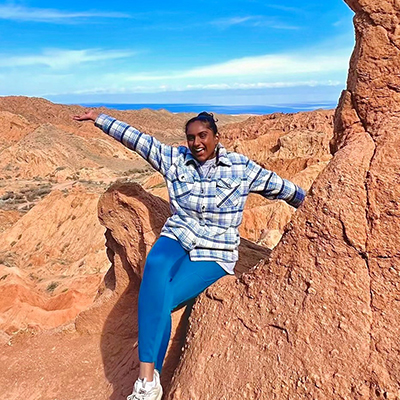 A woman in a plaid shirt and blue slacks happily waves from a red rock in a mountainous area.