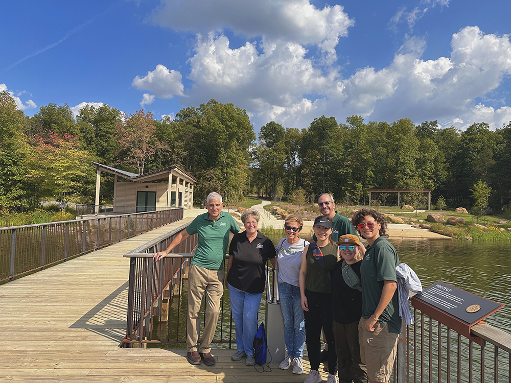 Recreation Management faculty pose with Trimner and Gilbert on a dock in Reservoir Park beneath blue skies and green trees.