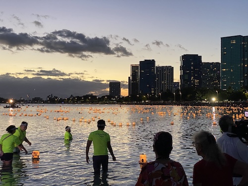 Lit floating lanterns filling up a bay in Hawaii with people standing in the water launching more lanterns
