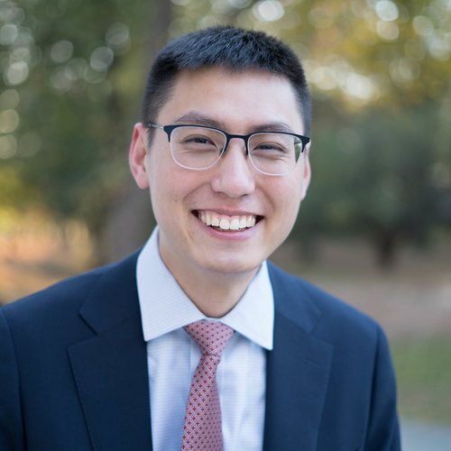 A man in a light blue collared shirt, tie, and dark blue jacket stands outside and smiles