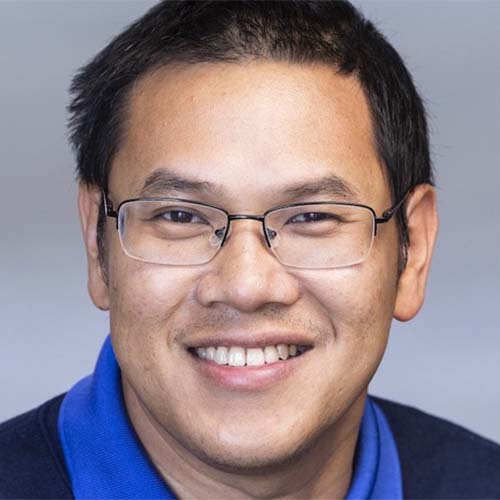 A headshot of CS associate professor ThanhVu Nguyen, wearing glasses and a blue collared shirt against a light gray background.