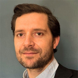 A man with a beard wearing a black blazer and white collared shirt stands and smiles