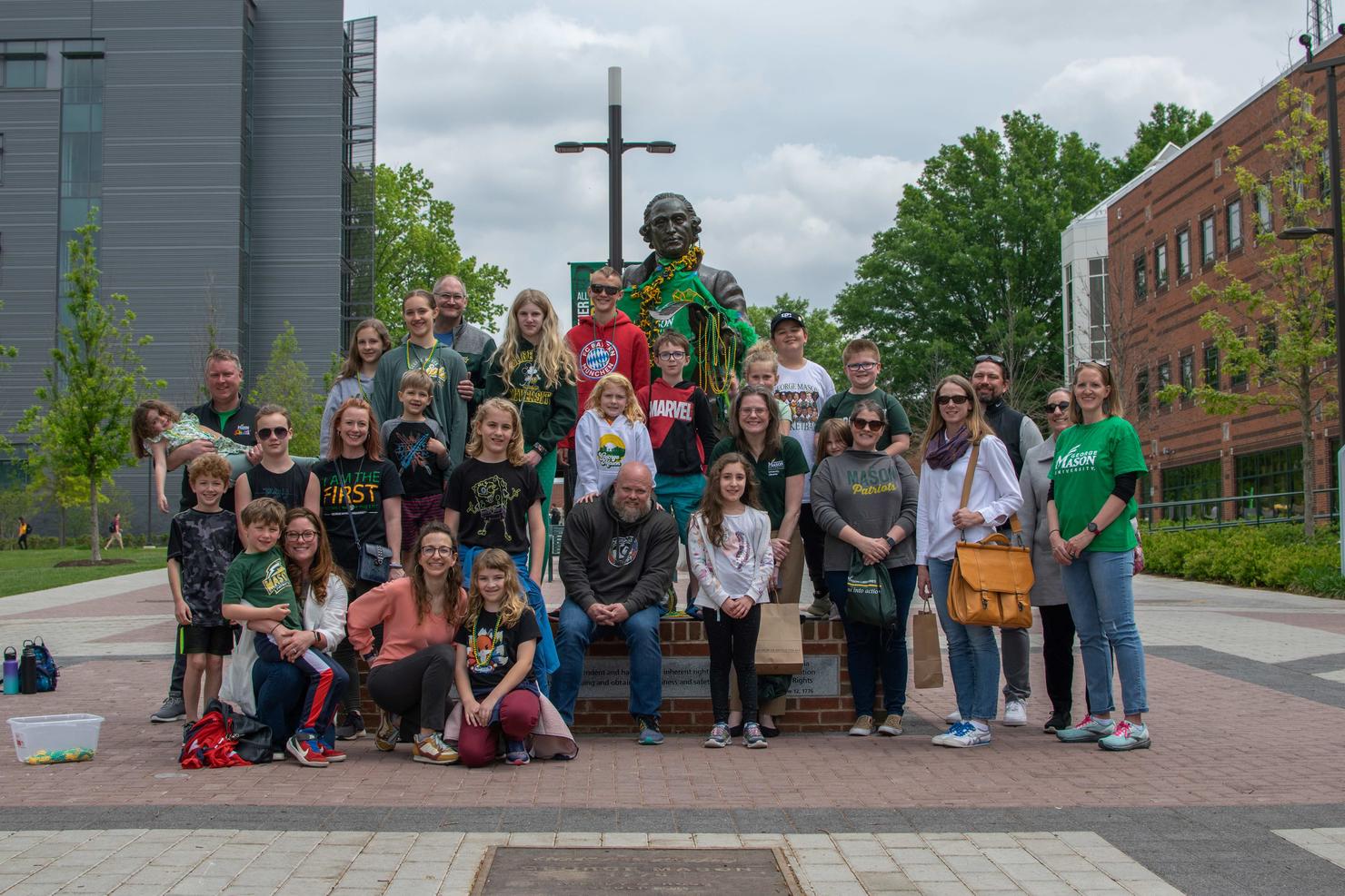 A group of about 30 employees and their children stand together at the George Mason statue on Take Your Junior Patriot to Work Day.