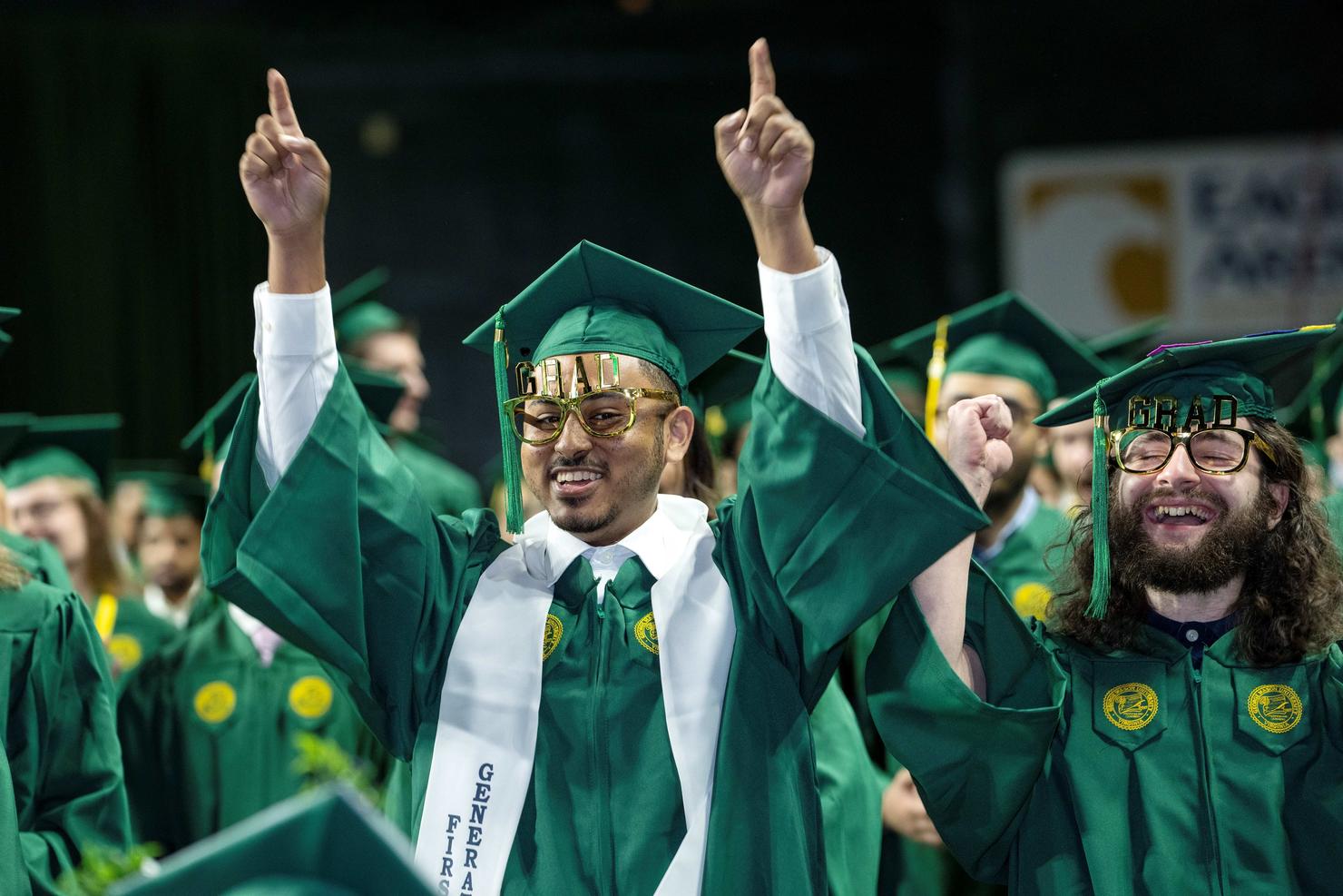 Graduate holds his arms up in victory while wearing glasses that say "Grad"