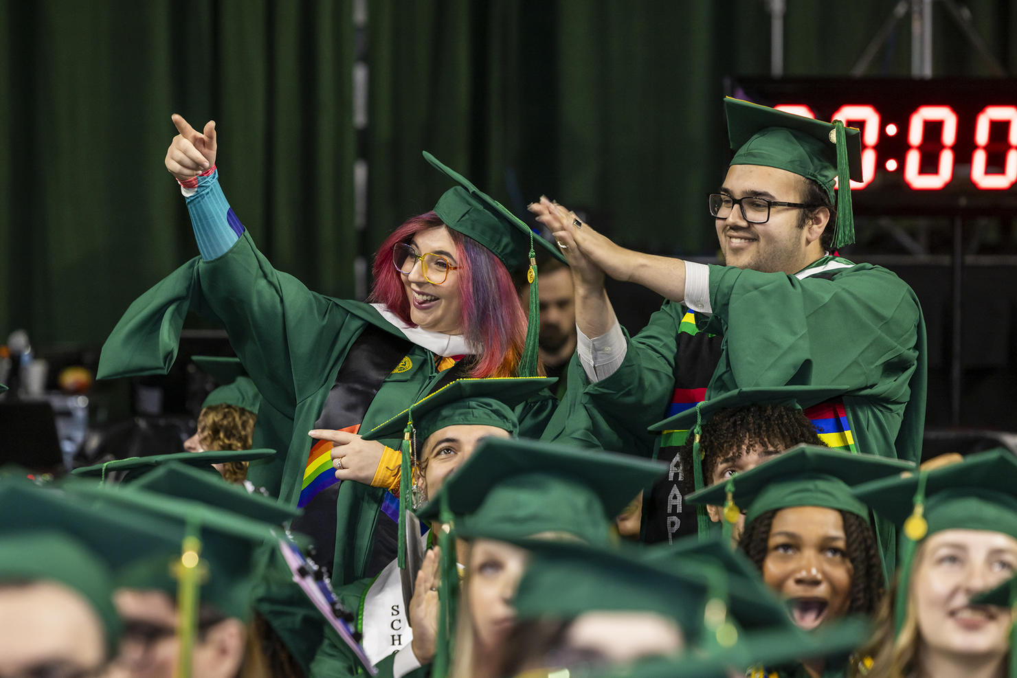 A woman in regalia with a rainbow stole and a man in regalia are standing up in the crowd at Spring 2024 Commencement
