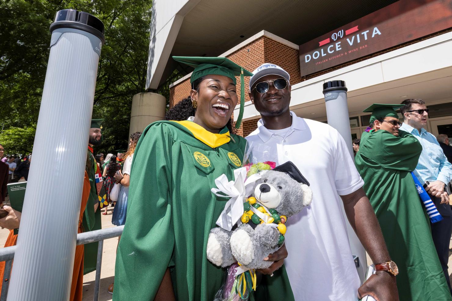 Graduate smiles with a companion while holding a teddy bear outside EagleBank Arena