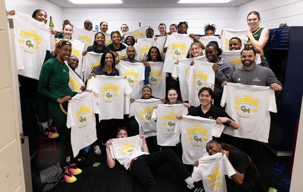 The women's basketball team and coaches hold up commemorative t-shirts, grinning in their team locker room.