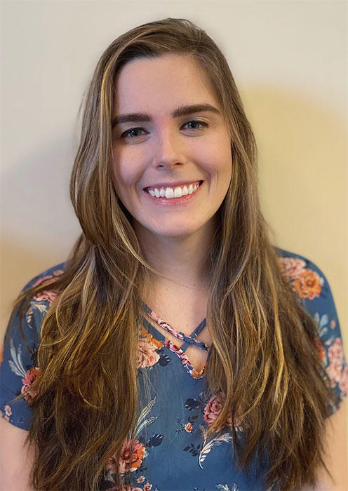 Kelly standing in front of a beige wall facing the camera, smiling. 