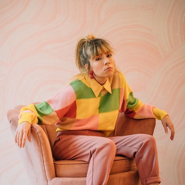 A young woman, Jeannette McCurdy, sits on a pink-peach color chair. She is wearing a block colored blouse with yellow, green, peach, and peach colored pants.