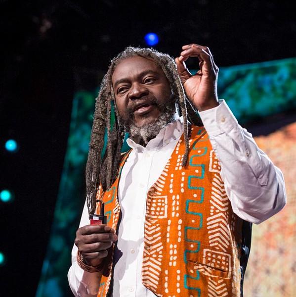 Visual Voices Lecture Series with Saki Mafundikwa. Saki is a Black man with long dreadlocks, he is wearing a white button down shirt and an orange African print vest.