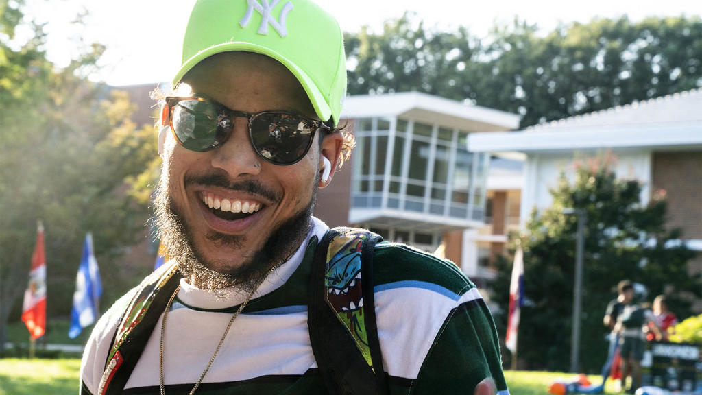 a young Black man smiling wide, wearing sunglasses and a green hat poses for the camera