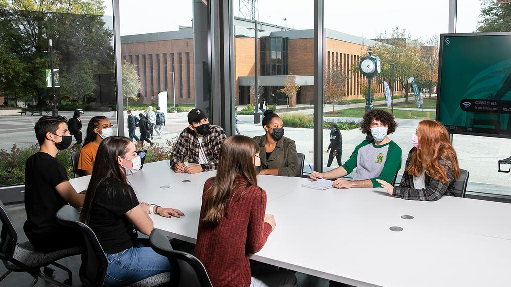 A group of diverse students sit at a conference table in a room where the walls are made of windows. These windows look out onto more students walking outside.