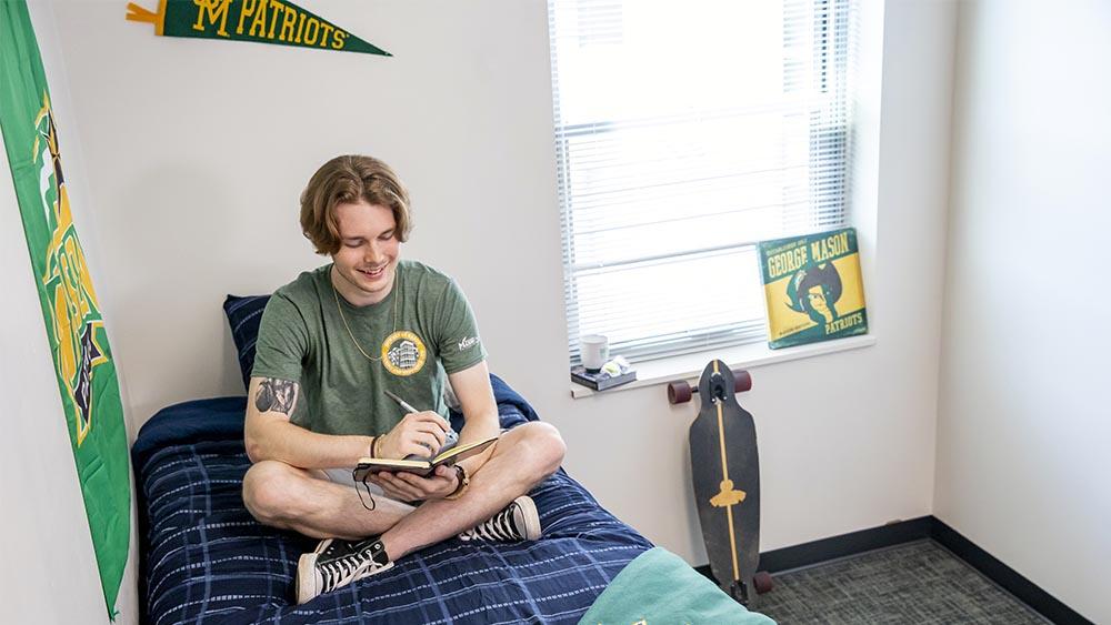 a young male student sits on his bed in a single dorm room. A skateboard leans against his wall.