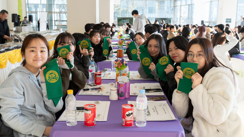 Mason Korea students sit around a long rectangular table, holding up 2024 New Student Convocation banners.