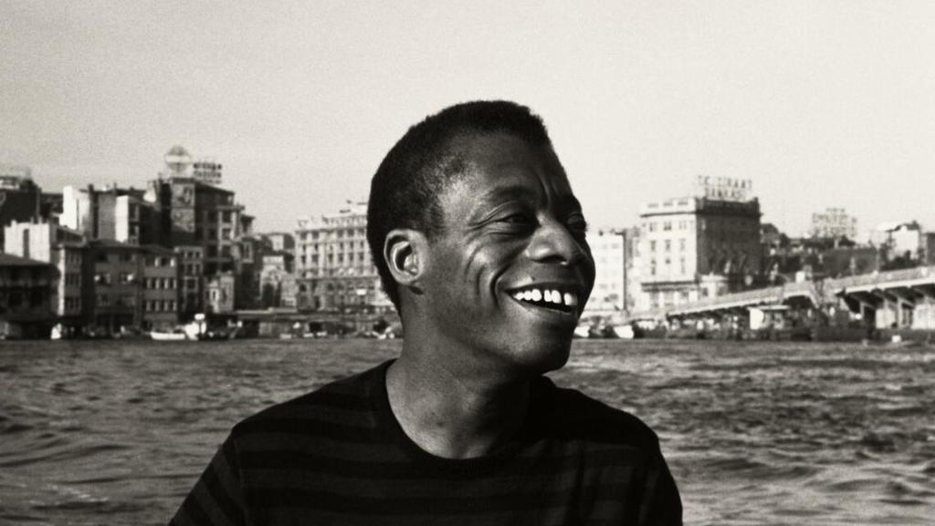 A black and white 1966 photograph of author James Baldwin during the time he lived in Turkey. A body of water and the city in the background.