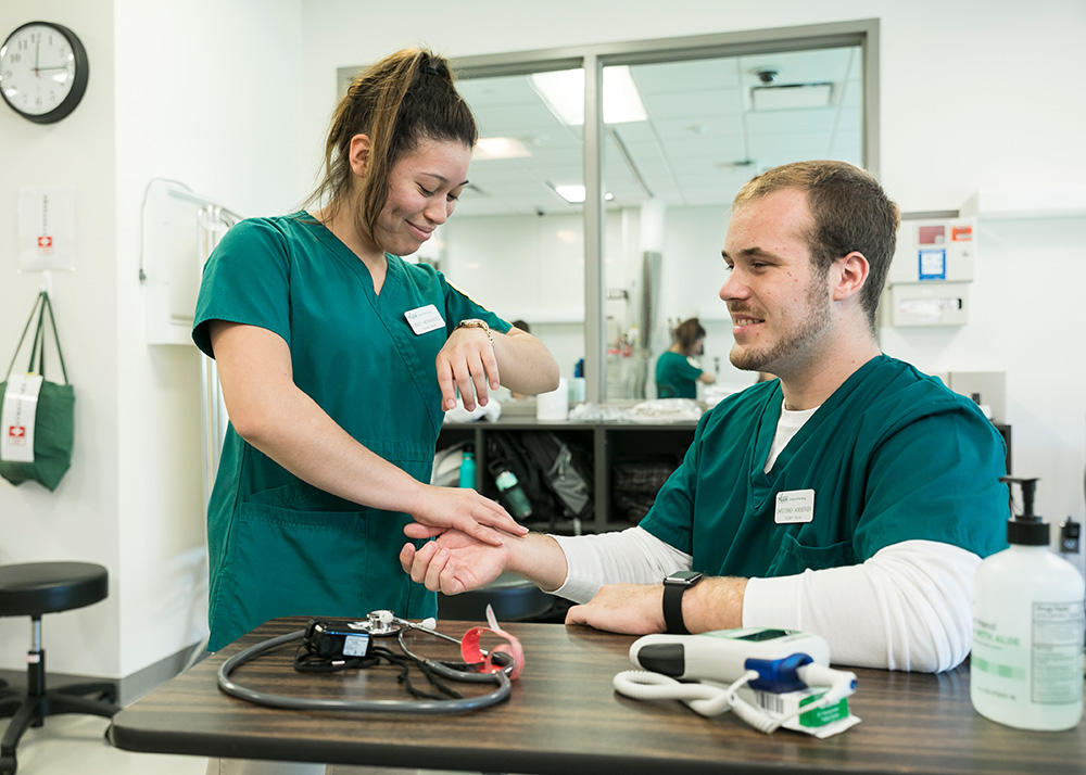 A student nurse checks the pulse of another student nurse in a practice scenario.