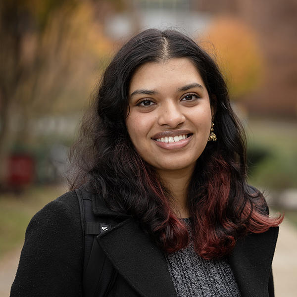 Female student Daksha Magesh looks at the camera for a headshot taken outside. She wears a coat and sweater and has a big smile. 