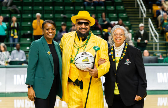 Doc Nix Nickens, Green Machine Ensembles director dressed in a bright yellow suit, yellow hat, green mardi gras beads holds a basketball. He is flanked to his right by interim athletic director Nena Rogers, wearing a green blazer, and his mom to his left, wearing a black suit.