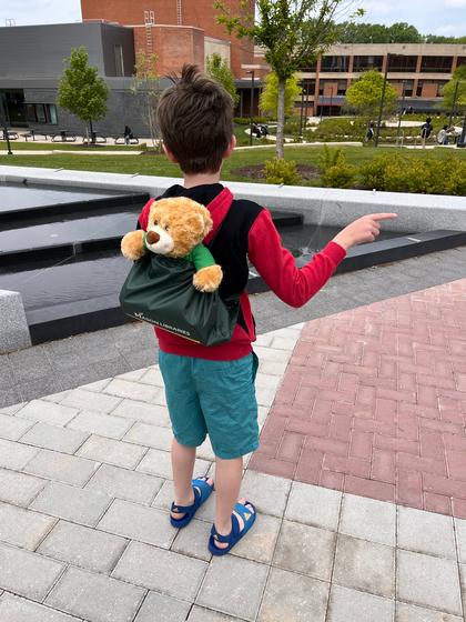 A child stands on the Fairfax Campus near the fountain on Wilkins Plaza. He has a teddy bear sticking out of his backpack.