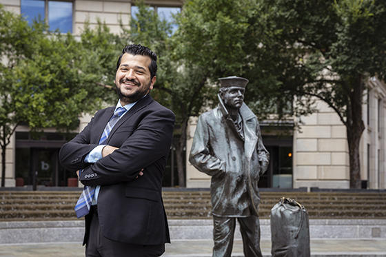 Marcou Shodi is in a navy blue suit, standing with crossed arms and smiling for the camera in front of a bronze statue of a sailor.