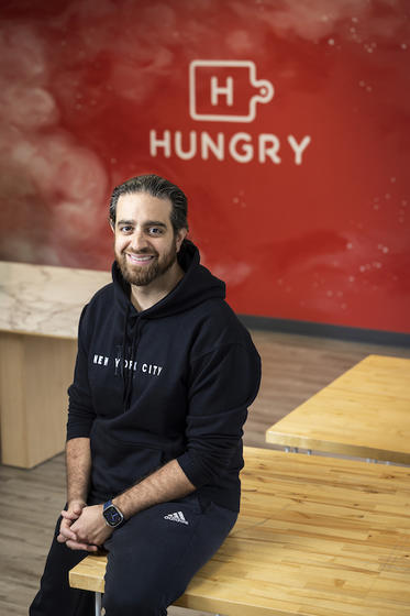 Mason alum Eman Pahlavani sits on a worktable in front of a red display wall with his company name Hungry