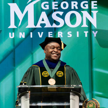 Gregory Washington, George Mason University president, stands behind the podium in Eaglebank Arena, dressed in full doctoral regalia