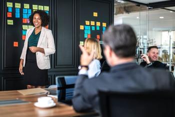 Image of several adults in business attire with a board with sticky notes, laughing and talking/brainstorming.