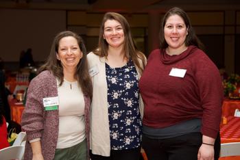 Three Mason employees smile together at the camera. Round tables with orange tablecloths and flower arrangements are in the background