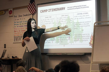 A teacher stands at the front of a classroom of students. She motions to text being projected onto a screen behind her, addressing the class.