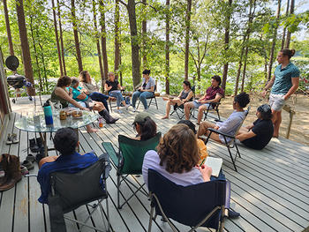 Students sit in camping chairs on a wooden deck, surrounded by green trees. 