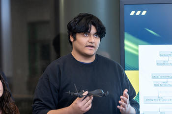 A student with black hair and tan skin holds a prototype of a drone in one hand and speaks emphatically with the other.
