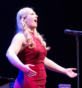 A woman with long blonde hair in a formal red dress sings for a crowd.