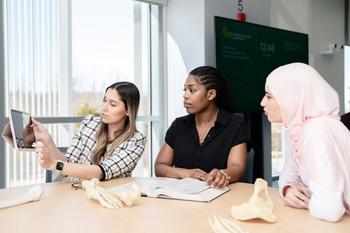 Three science students work together in a classroom, with one student showing a tablet to the others. Model skeleton pieces are on the tabletop