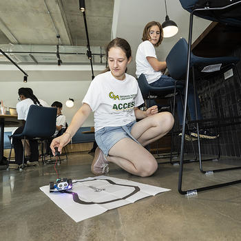 A student kneels by the paper track with a micro:bit robot. Another student behind her watches.