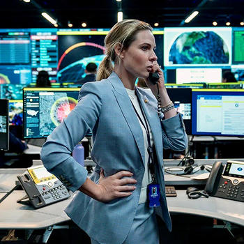 A woman in a blue suit stands amid a lot of computer screens and talks on a telephone.