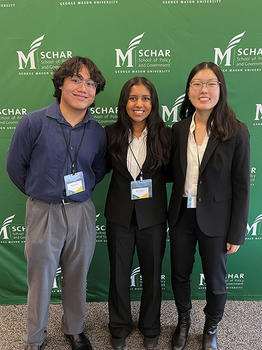 A group of three students stand in front of a green banner with the Schar School logo