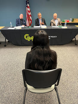 A woman with long black hair sits in a chair facing three men sitting at a table with an American flag behind them.