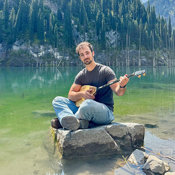 A man with dark hair and a short beard plays a two-stringed instrument while sitting on a rock in a shallow lake with mountains behind him.